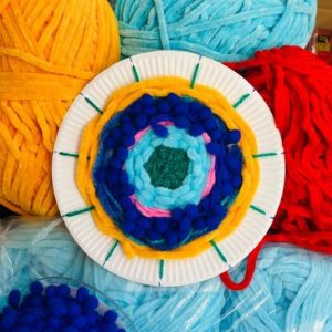 A colour photograph of a close up of balls of wool on top of which is a round paper plate that has the beginnings of a circular wool weaving