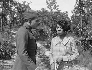 A mono still from the film The Flying Ace of a young man and woman. He is dressed in army uniform, she in a collared dress. He is looking towards her and she is looking sadly towards the ground.