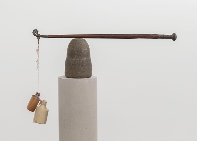 A sculpture of a concrete column upon which is a dark brown, cone shape, and then balanced on top is a long pole with a hook at the left end. Hanging from the pole, are two conical ceramic containers, one cream and one brown.