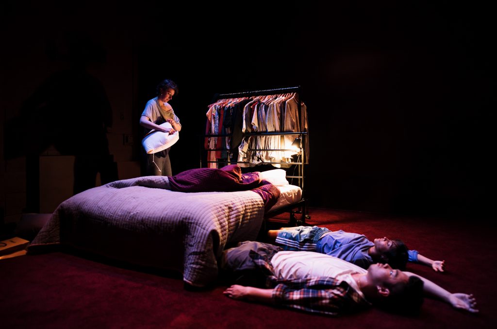 A woman wearing a grey t-shirt an leggings changes the sheets on a double bed, At the foot of the bed stands a crowded clothes rail, lit up by a dim lamp. In the foreground of the image on the opposite side from the woman, two people wearing similar casual dress lie flat on the thick red carpet, half-under the bed. One has their eyes closed and the other is staring at the ceiling. Both have one arm raised above their head.