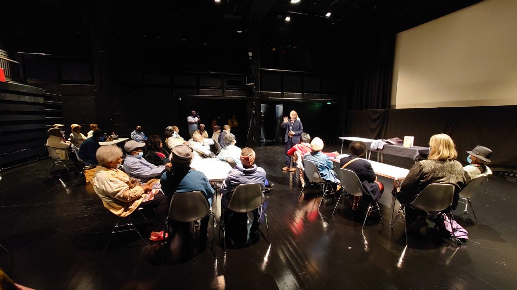 IN the Arnolfini Theatre, a dark room with spotlit tables and chairs, A group of people from various caribbean elders community groups sit listening to Gary Topp, Director of Arnolfini share the work that they do.