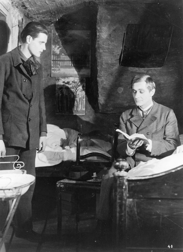 A black and white image from the film Phantom, of Alfred Abel seated on a bed, reading a book, while Hans Heinrich von Twardowski opposite.