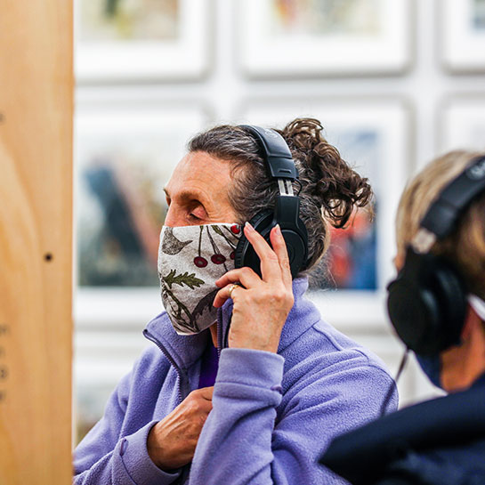 A colour photograph of a person wearing audio headphones, which they are holding to their ear, within a Gallery at Arnolfini