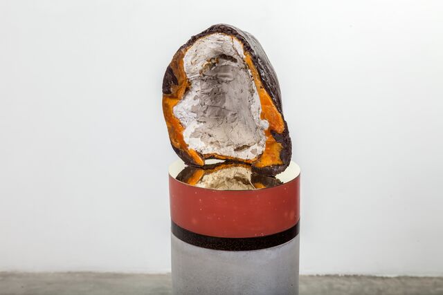 An abstract sculpture created in wax, concrete, plaster, and hessian fibre. It is oval in shape and textured, hollow with an outer dark grey, layer of dark orange-yellow and inner of white. It sits on a circle brass topped plinth.