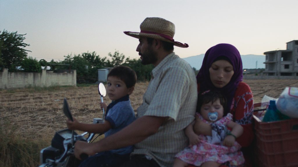 Wild Relatives, 2018 (Walid, organic farmer) Jumana Manna A still image taken from a film called Wild Relatives made by artist and film-maker Jumana Manna. The image is of three people on the back of a small motorbike. A young boy is holding the side mirrors at the front and a man wearing a wide brimmed straw coloured hat sits behind holding the handle bars. Behind him sits a woman in a headdress holding a small child. In the background is a dusty landscape with a dirty white wall and trees on the left and a two-storey building on the right.