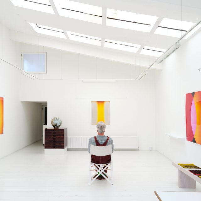 A colour photograph of artist Garry Fabian Miller sat in a white walled and floor studio, surrounded by his work.