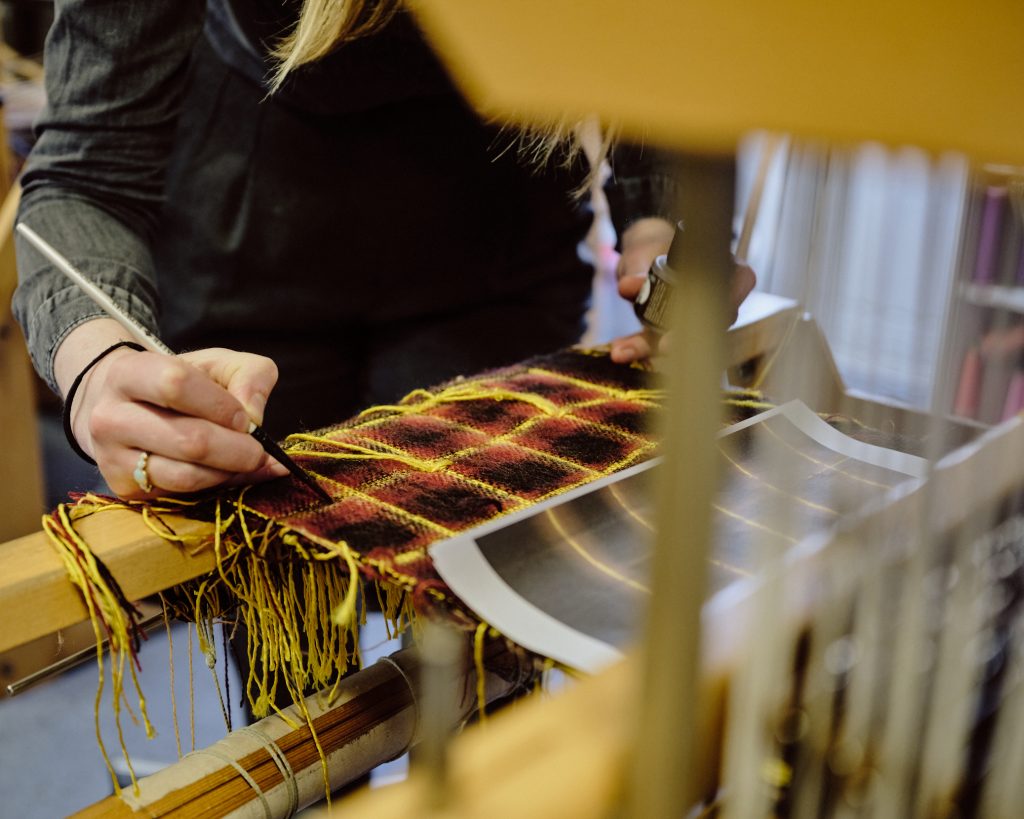 A close up colour photography of a weavers loom at which someone is hand painting a piece of tapestry in front of them.