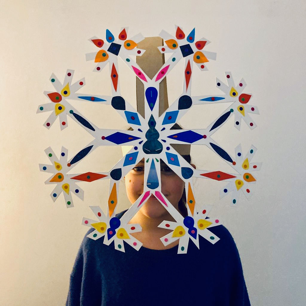 a young girl from the chest up stands facing us wearing a large paper snowflake mask that obscures her face. The white paper snowflake is decorated all over with different coloured shapes.