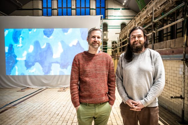 A photograph of artist duo Juneau Projects, Phil and Ben, standing smiling in a disused swimming pool.