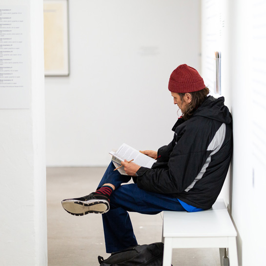 A colour photograph of someone sat, on a bench, in one of the galleries at Arnolfini