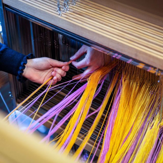 A pair of hands touching yellow and pink thread on a loom.