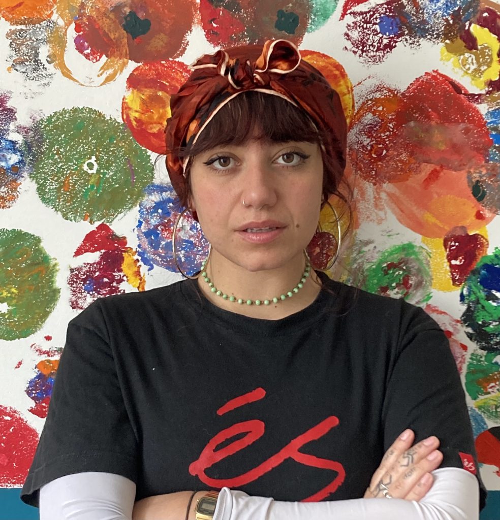 A photograph of the artist Iman Sultan West looking into camera with crossed arms and wearing a black t shirt against a background of splodged paint.
