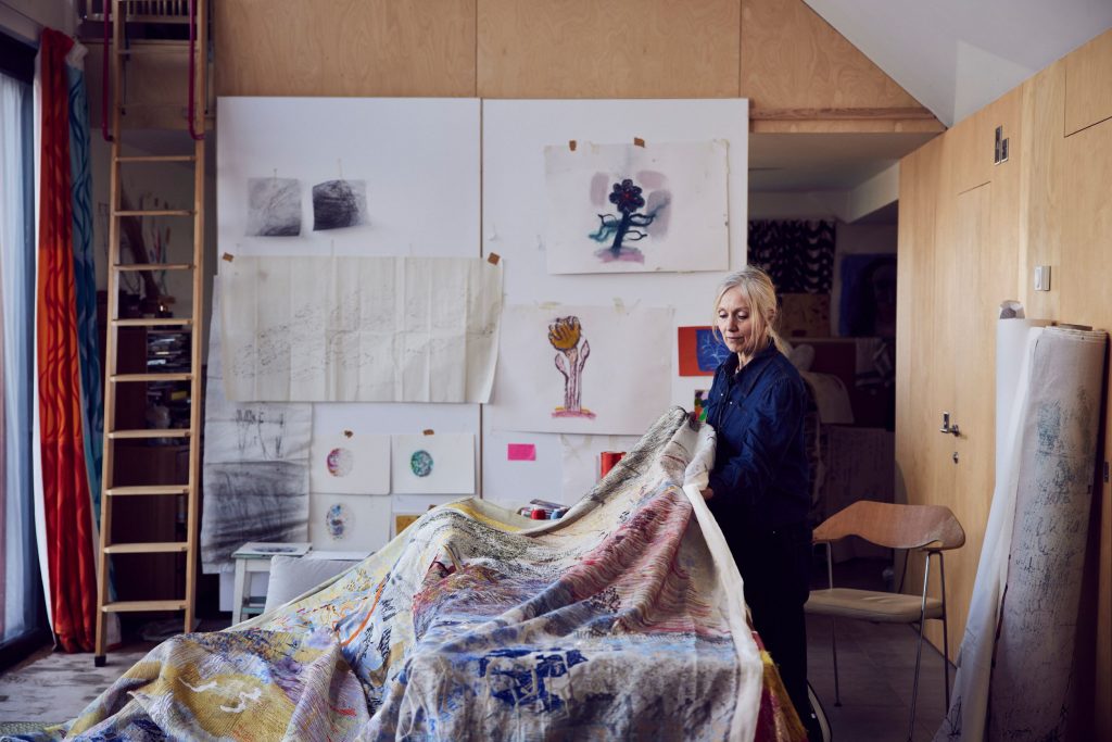 Alice Kettle stands in her studio space holding the corner of one her large-scale textile works which drapes over a table in front of her. The surface of the work is complex and textured with colours including yellow blue and red. Behind her, a number of sketches are pinned to a wall.