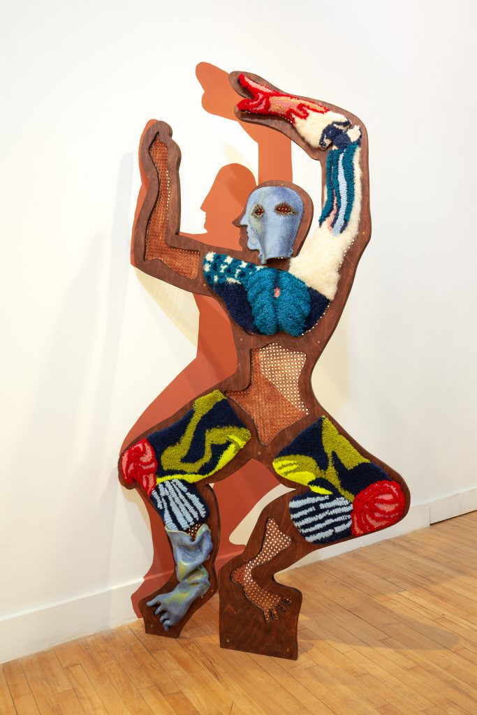 A colour photograph of an artwork of a human figure, arms in the air, squatting slightly. It is made from a mixture of materials with the main body wood and wooden mesh and parts of the body in wools and material.