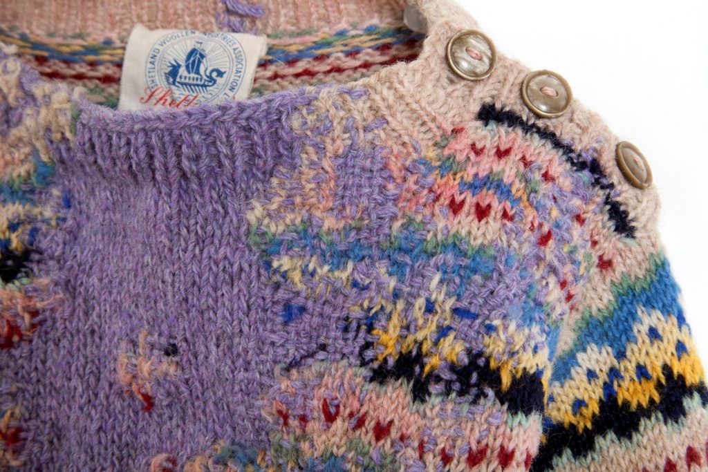 A knitted jumper that has been visibly mended by artist Celia Pym. The jumper features a geometric pattern in various shades of blue, red, cream and black. Various aspects have been visibly mended in purple wool stitches, creating a deliberate visual contrast with the original pattern of the jumper.