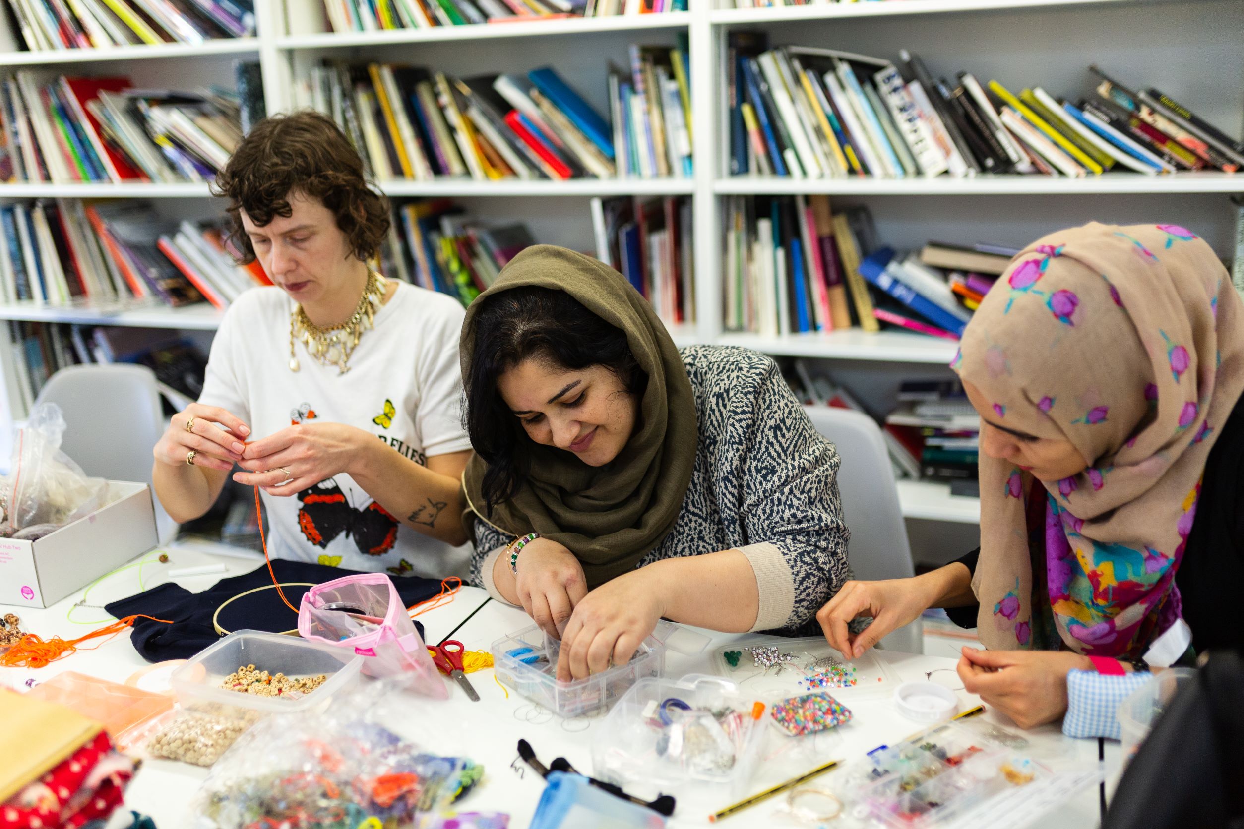 A group of people gathered around a table in an art studio, creating with different materials including beads and threads. A shelf with art supplies is in the background. The mood is one of creativity, community, and collaboration.