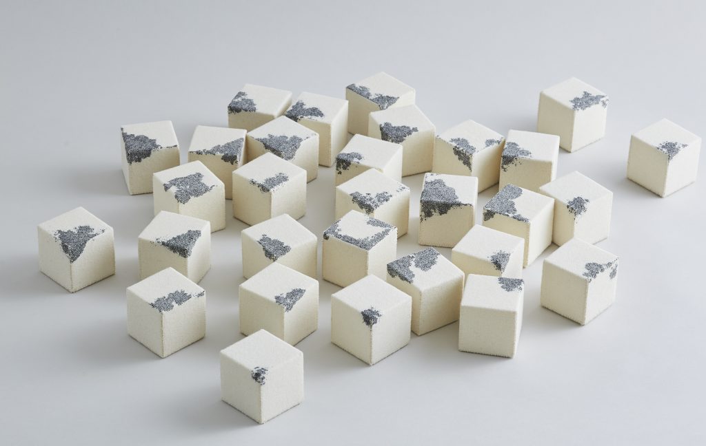 A colour photograph of Richard McVetis' work entitled variations of a stitched cube, featuring a number of off white, fabric cubes, each with an abstract element stitched onto them in blue thread.