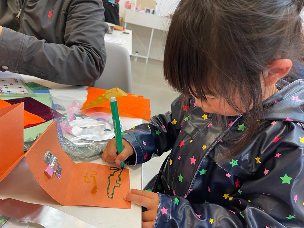 a young child is busy decorating an orange piece of card on the table in front of them. They are holding a green, felt tip pen with which they are drawing a shape. The table is adorned with a variety of creative materials.