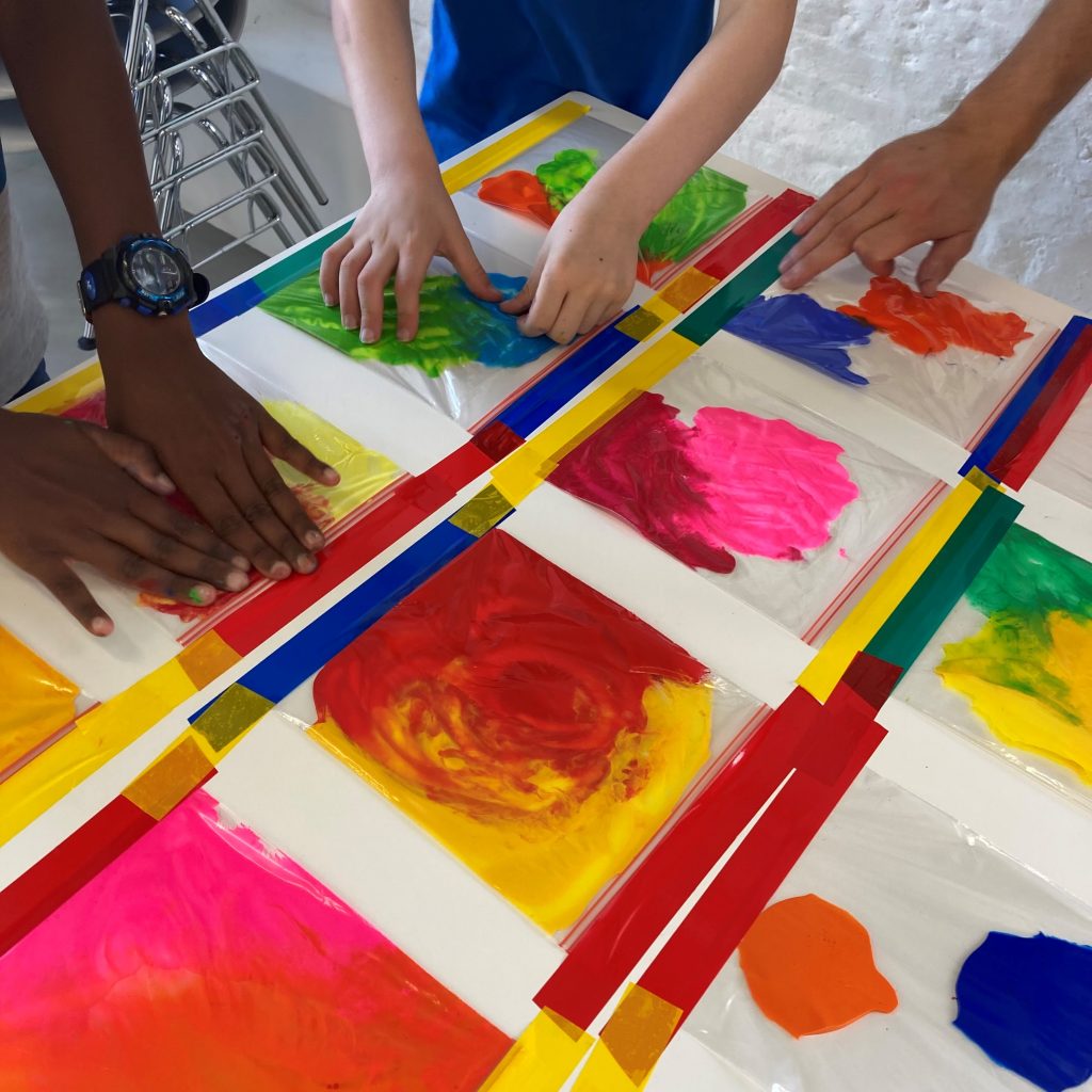 A table is laid out with lots of coloured squares spread with paint. Two pairs of hands are laid onto the squares of paint to smooth the paint around.