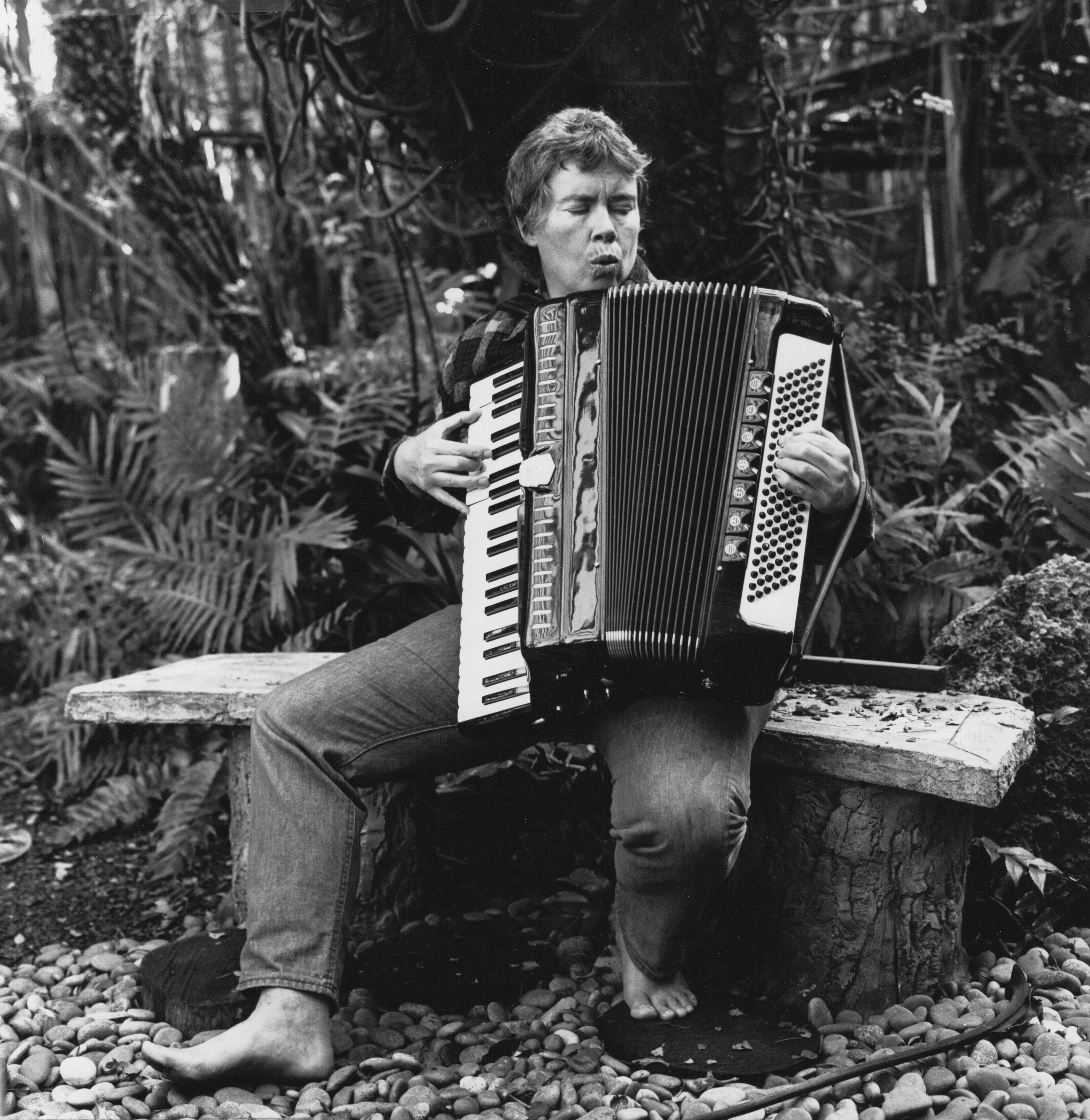 a black and white photograph of musician Pauline Oliveros, taken in 1979, seated on a stone bench, playing their accordion.