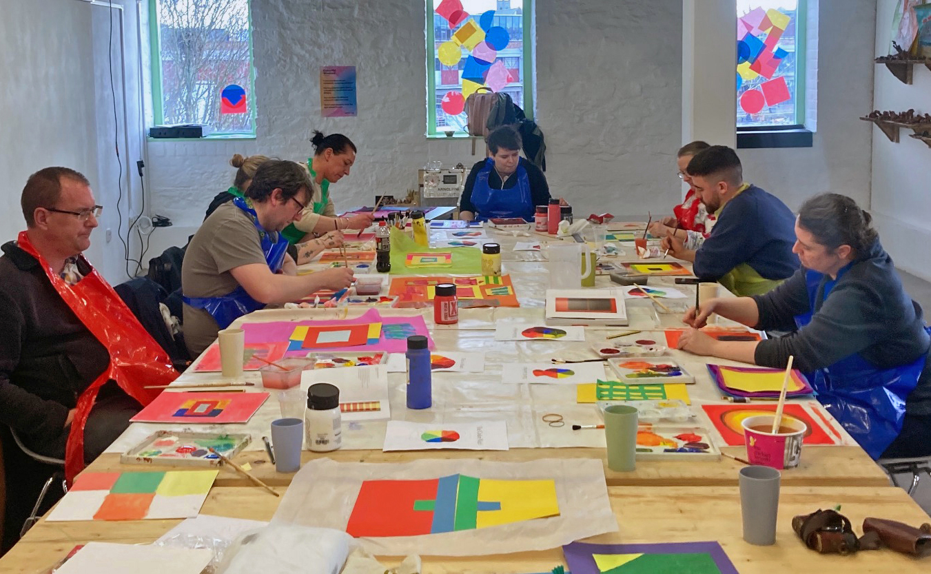 a group from AIM Art School busy creating with colour paper onto white paper laid out on a set of tables in the community workshop space at Arnolfini.