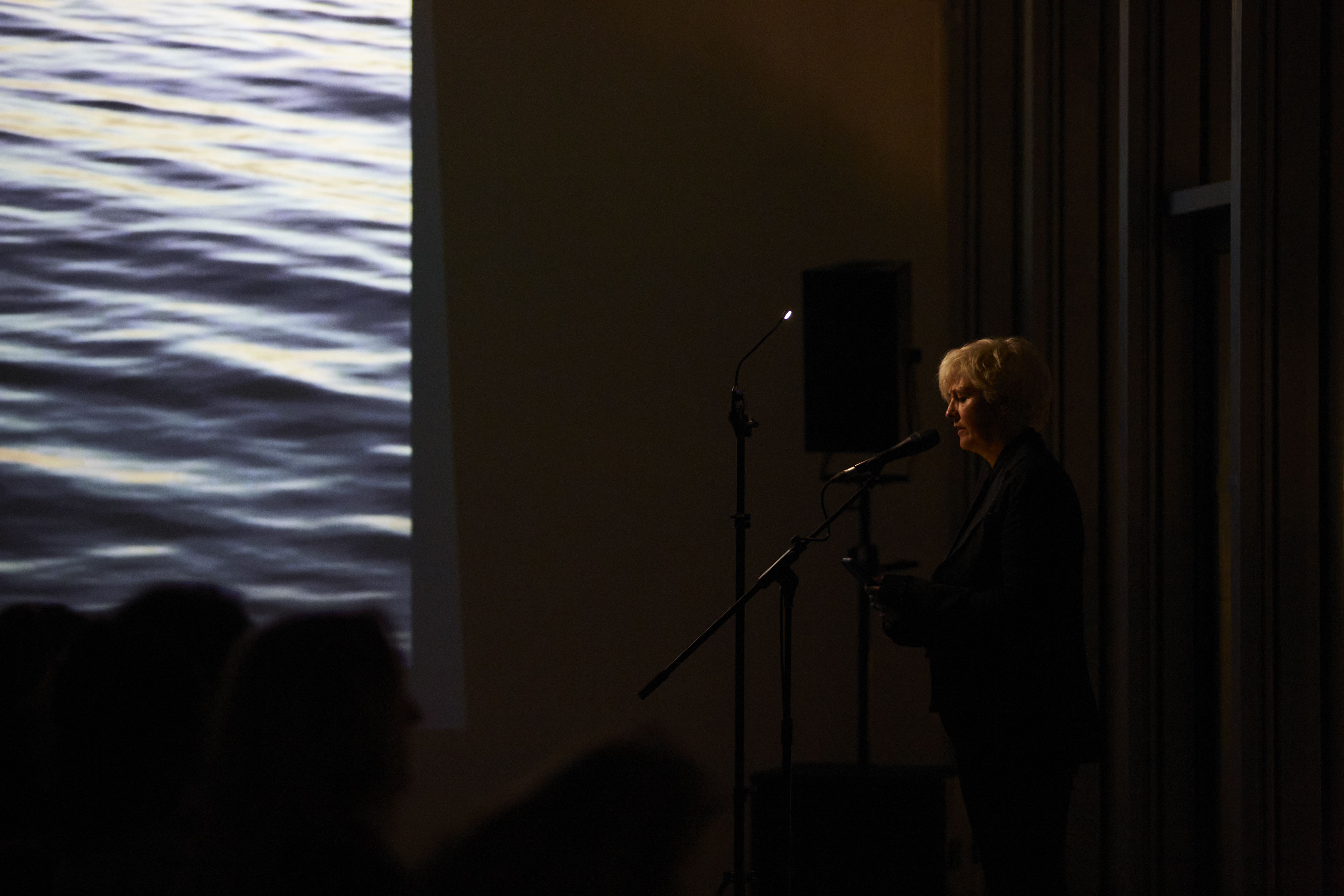 A colour photograph of artist Caroline Bergvall performing Nattsong. Caroline is sat next to a large screen on which is projected an image of water. the image is dark making Caroline almost a silhouette in appearance.