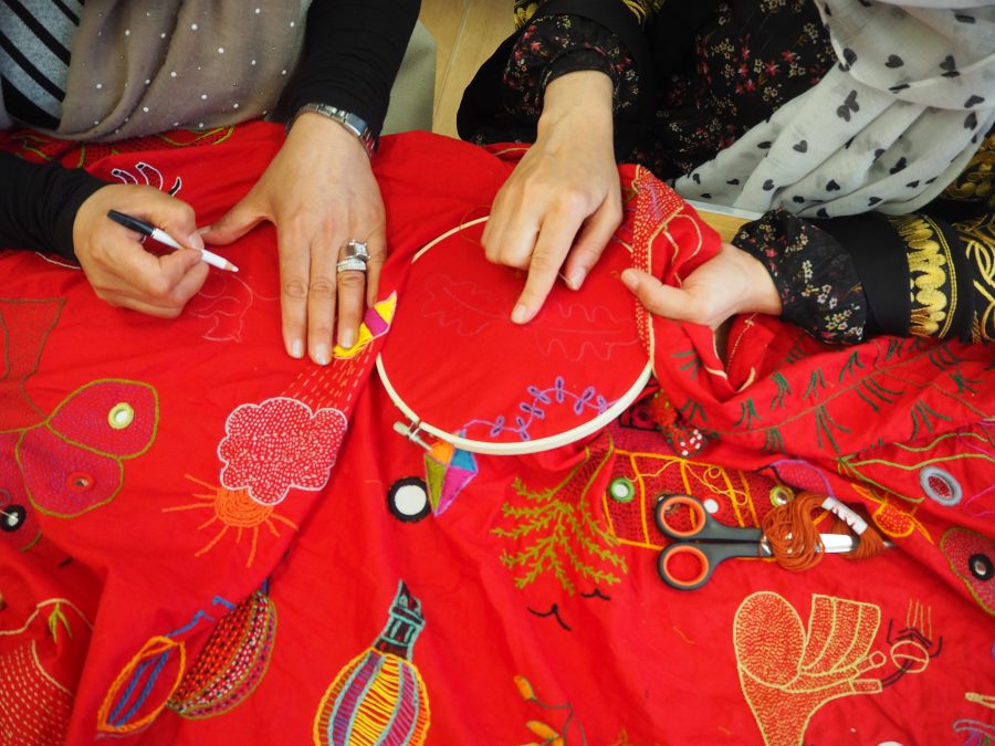 a colour photograph taken looking down onto a table on which are two set of hands working on an embroidered piece of red fabric.