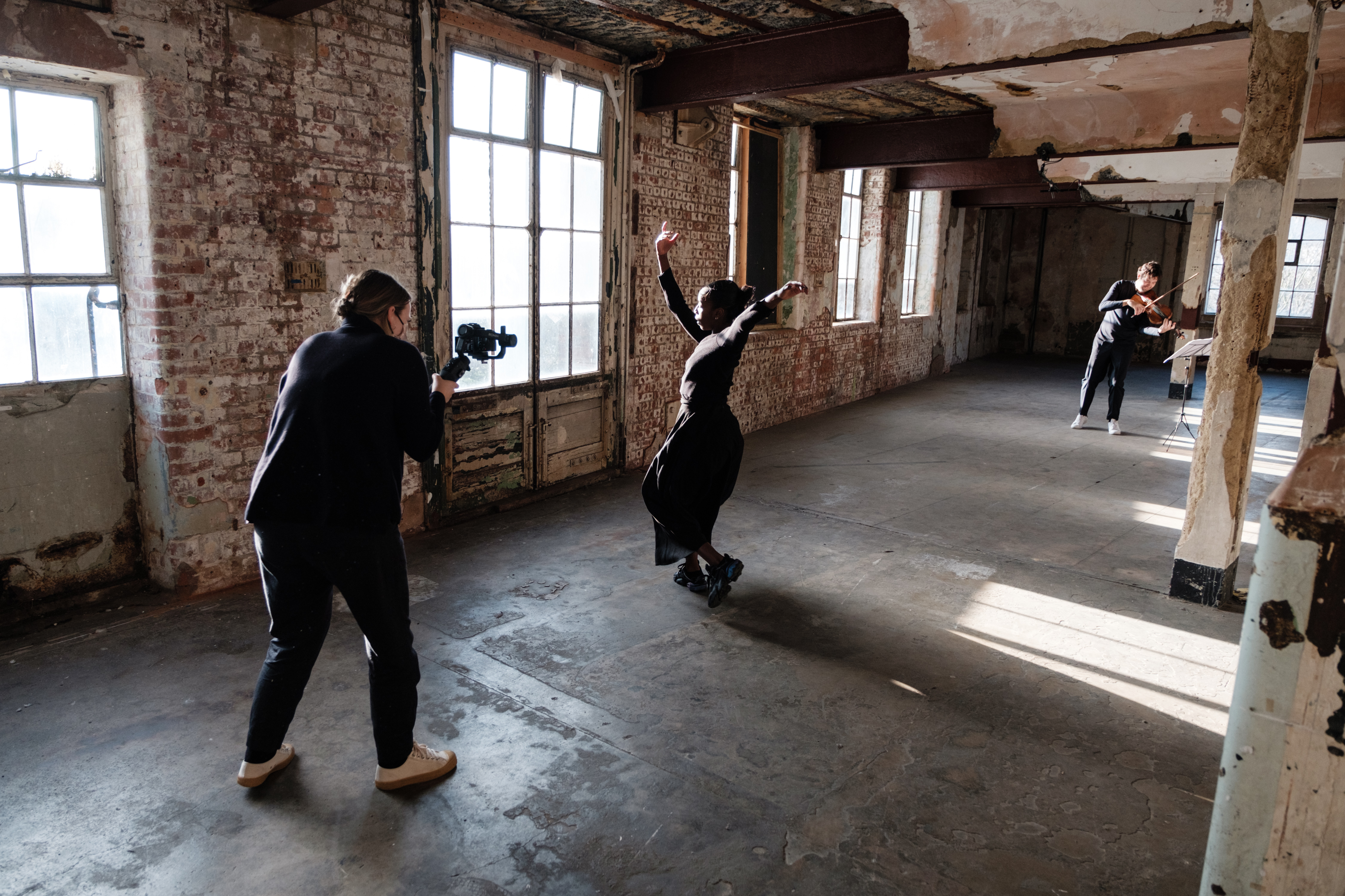 A colour photograph of performers being photographed in a large warehouse space. The photographer is closest in the scene, taking images of a dancer in the middle of the space and playing in the direction of the dancer, at the end of the room, is a seated violinist.
