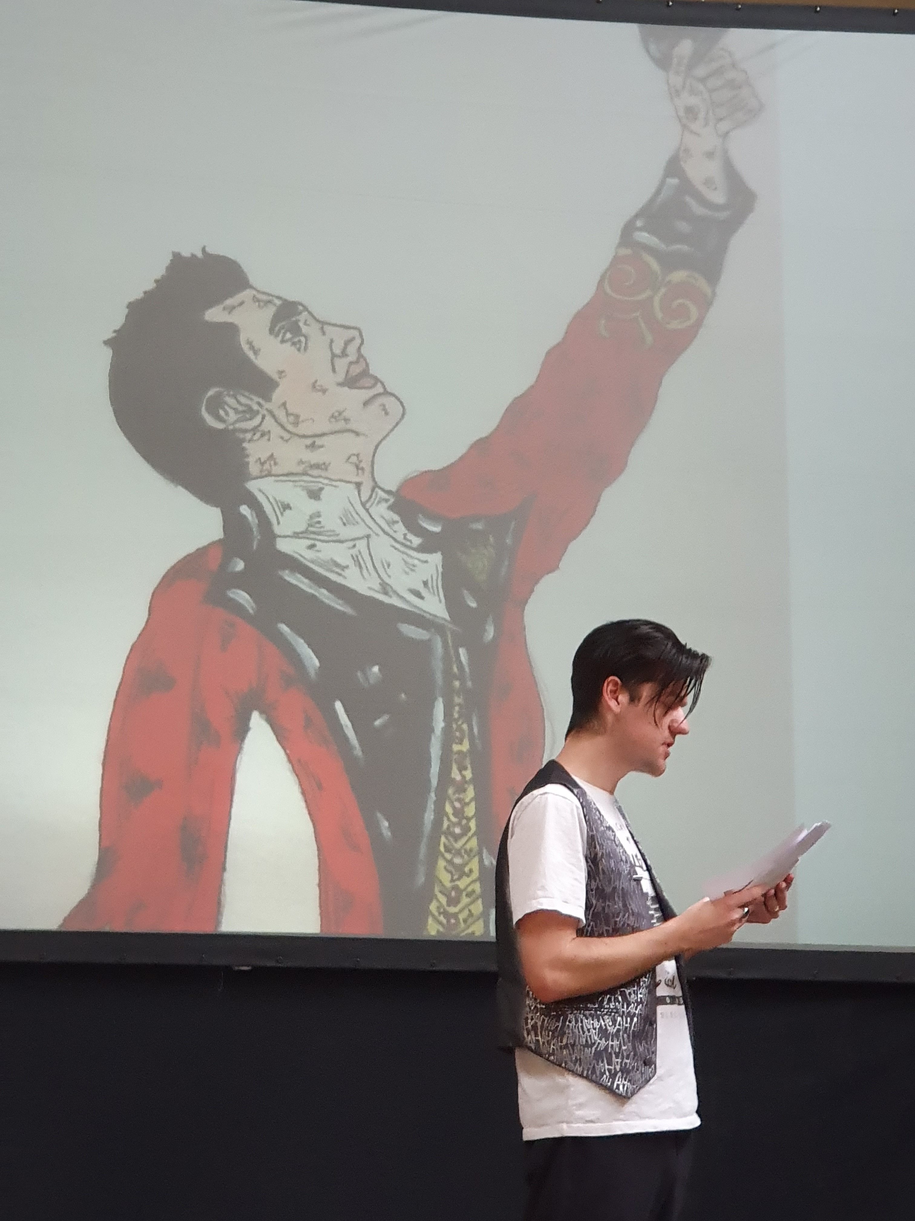 A coloured photograph of a man standing side profile reading from a piece of paper, perhaps addressing an audience that we cannot see. There is a large projection directly behind the man of an illustration of a man also in side profile.