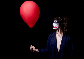 A young female performer wearing a dark blazer and clown makeup looks at a red balloon she is holding.