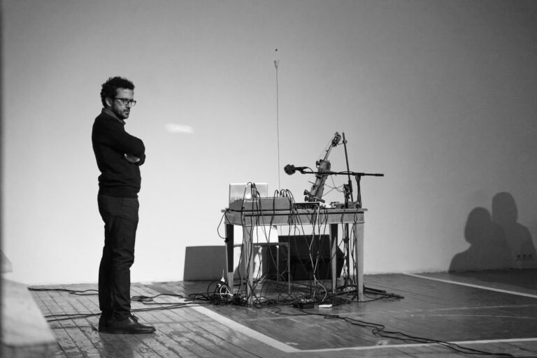 a black and white photograph of a person, wearing dark sweater and trousers, arms folded and looking towards the floor. In the same space, a table with an assortment of sound recording equipment.