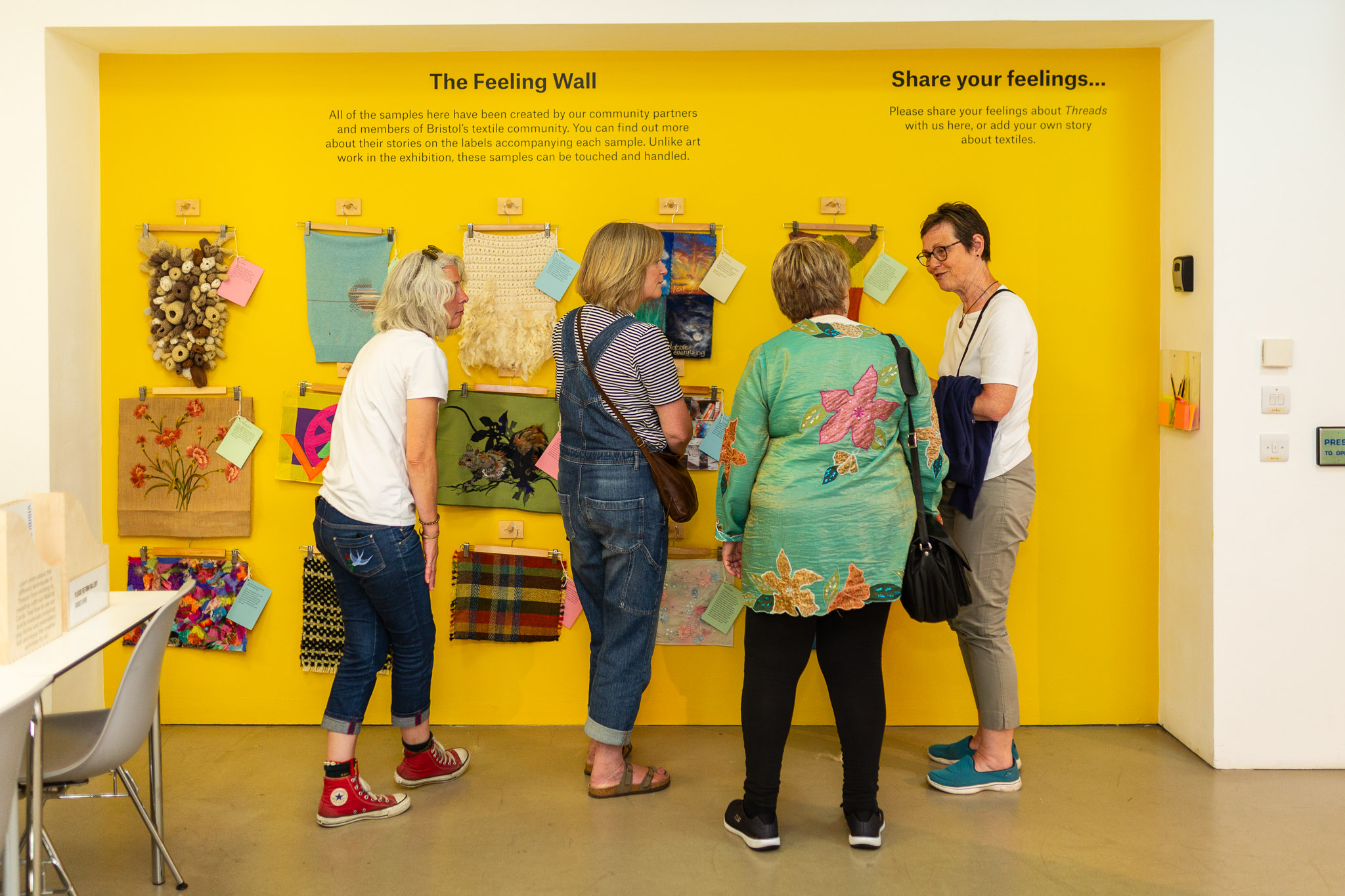 Four people are standing in a line talking to one another. They are facing a bright yellow wall on which are a collection of textiles.