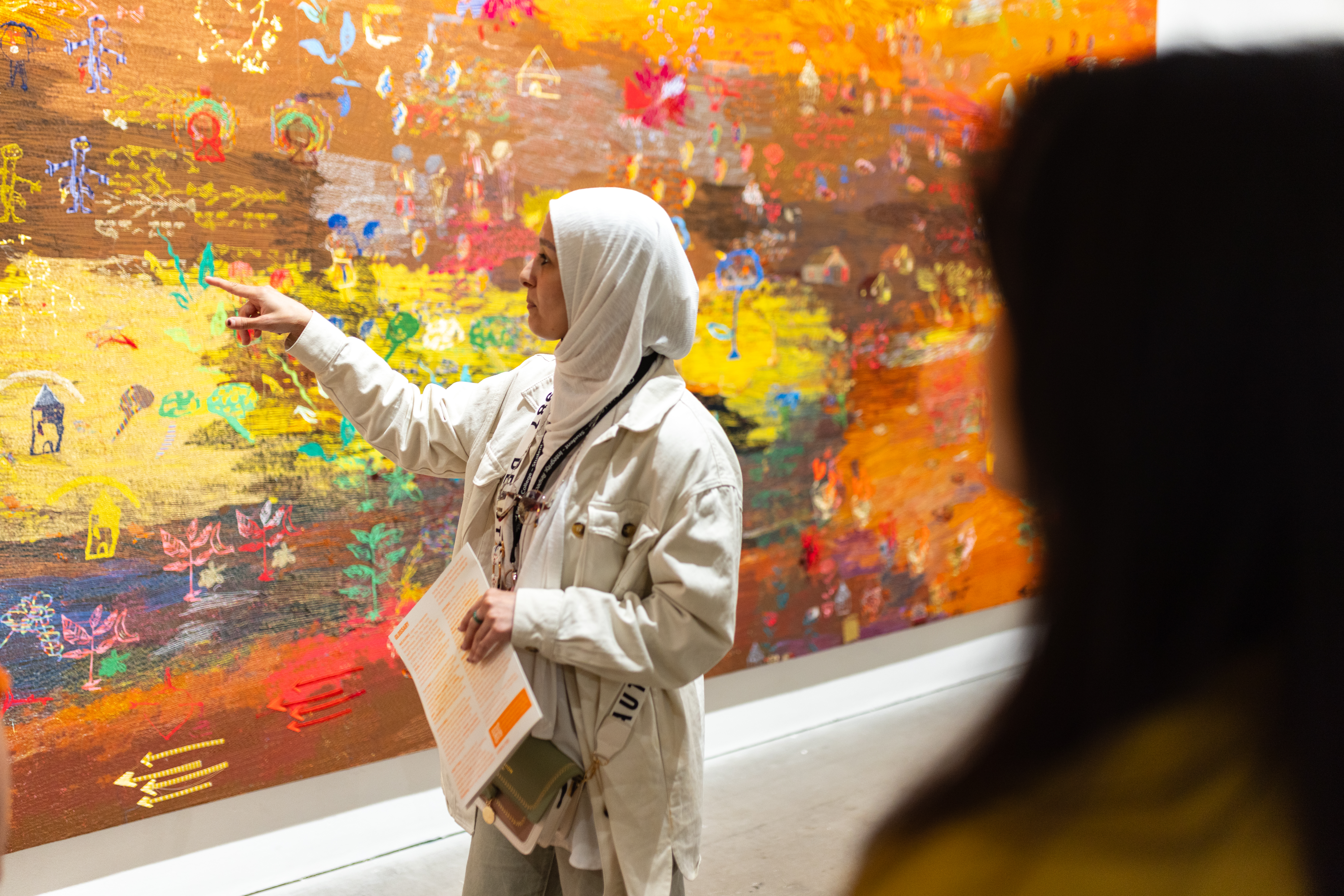 A woman dressed all in white with a white headscarf stands side on to a vibrant textile artwork on the wall. The textile work is a mixture of autumnal colours and heavily embroidered. She is pointing at the work.