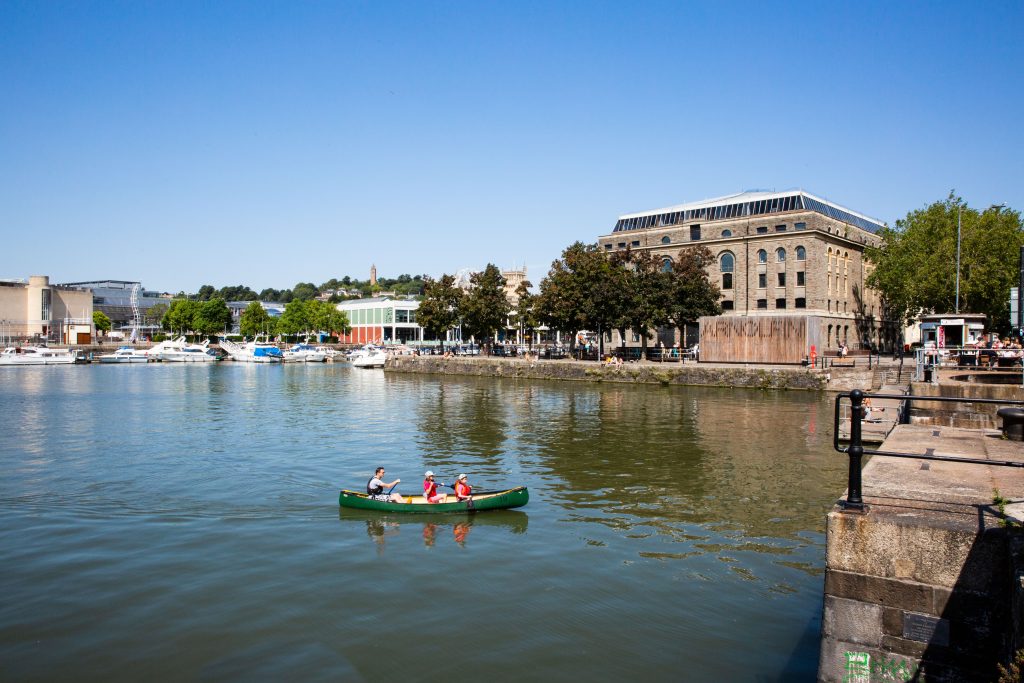 an exterior image of Arnolfini, from across the water. In the foreground, three people are travelling in a rowing boat.