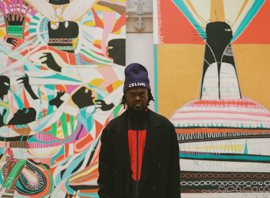 a photographic, colour portrait of artist Adébayo Bolaji stood in front of two of his works.