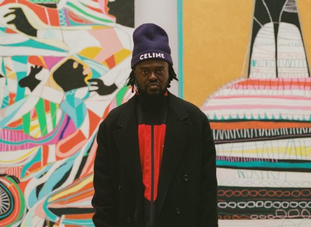 A colour photograph of artist Adébayo Bolaji stood in between his artworks No Beauty Without Struggle and The Vessel, both of which are 400cm in height. Adébayo Bolaji is dressed in a long, black coat over a peach coloured shirt and pale green trousers. He wears brown shoes and a dark blue, woolen hat with the word Celine stitched in white. He has just above shoulder length black hair and a beard and looks directly at the camera.