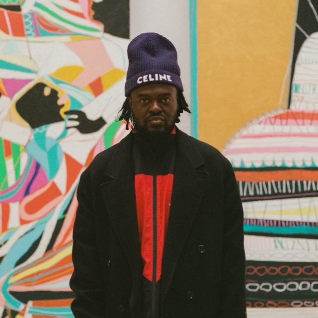 A colour photograph of artist Adébayo Bolaji stood in between his artworks No Beauty Without Struggle and The Vessel, both of which are 400cm in height. Adébayo Bolaji is dressed in a long, black coat over a peach coloured shirt and pale green trousers. He wears brown shoes and a dark blue, woolen hat with the word Celine stitched in white. He has just above shoulder length black hair and a beard and looks directly at the camera.